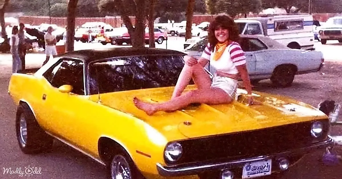 A woman sits on the hood of a bright yellow muscle car at a car show.
