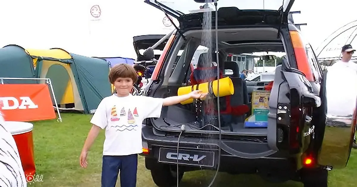 A boy stands next to an open car trunk, holding a yellow object as water pours from it.