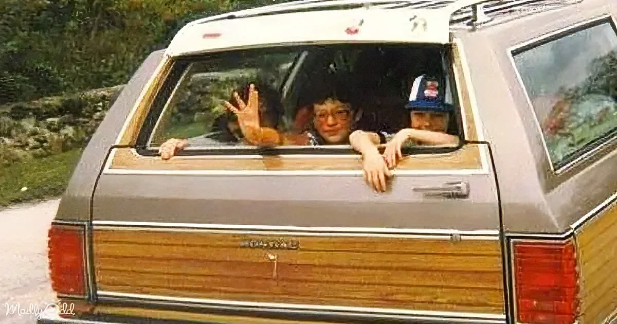 A vintage station wagon with wood paneling has two children leaning out of the back window, waving and smiling.