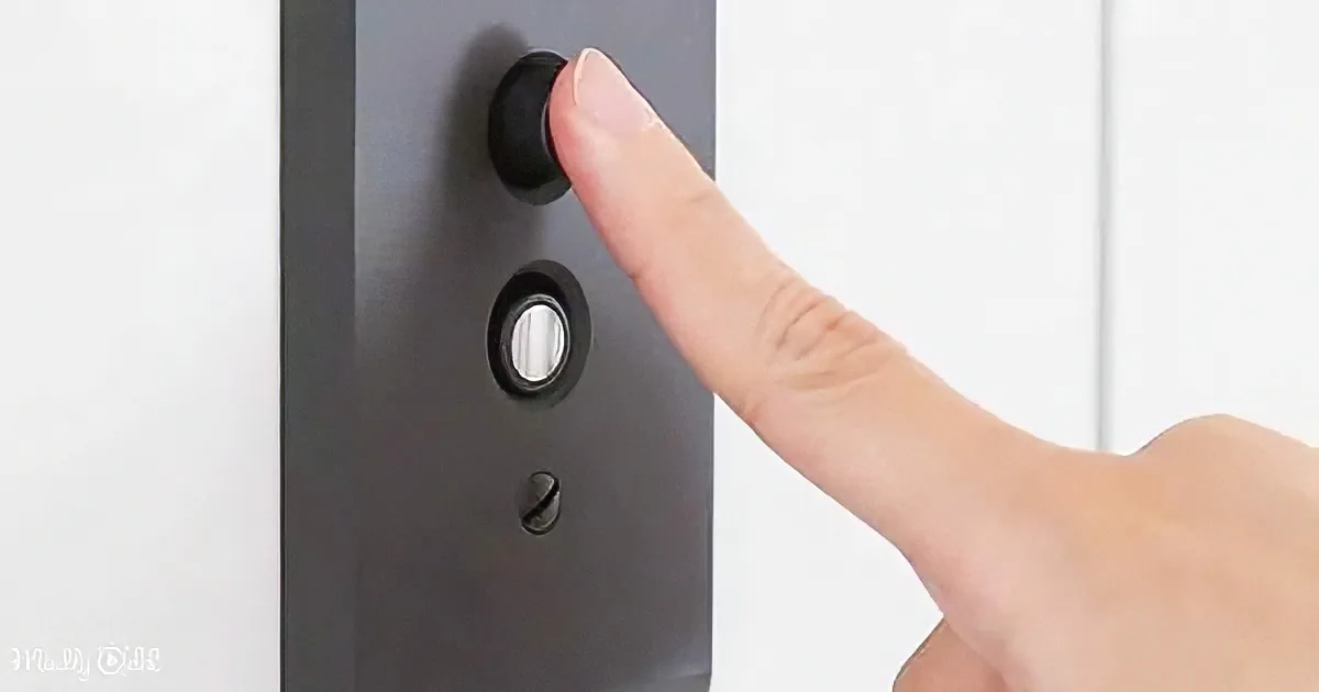 A hand presses a black button on a dark panel against a white wall.