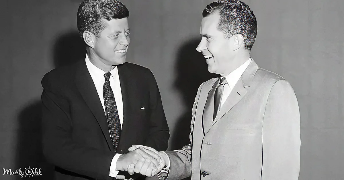 Two men in suits shake hands, both smiling, against a plain background.