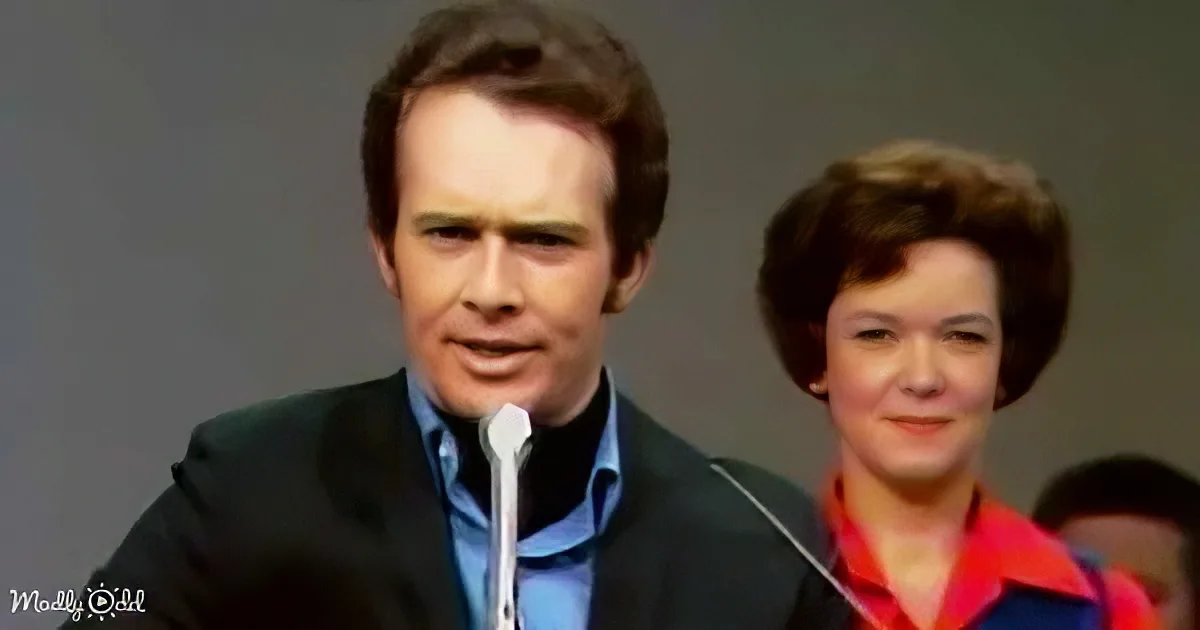A man in a suit speaks into a microphone, with a woman in a colorful outfit behind him.