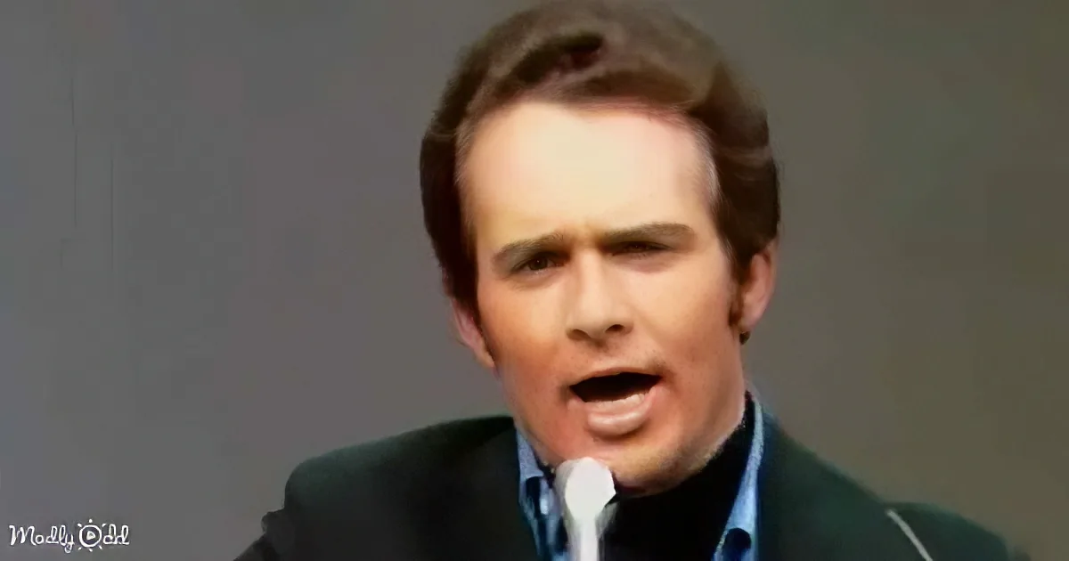A man in a suit and tie is speaking into a microphone, mouth open, against a plain background.