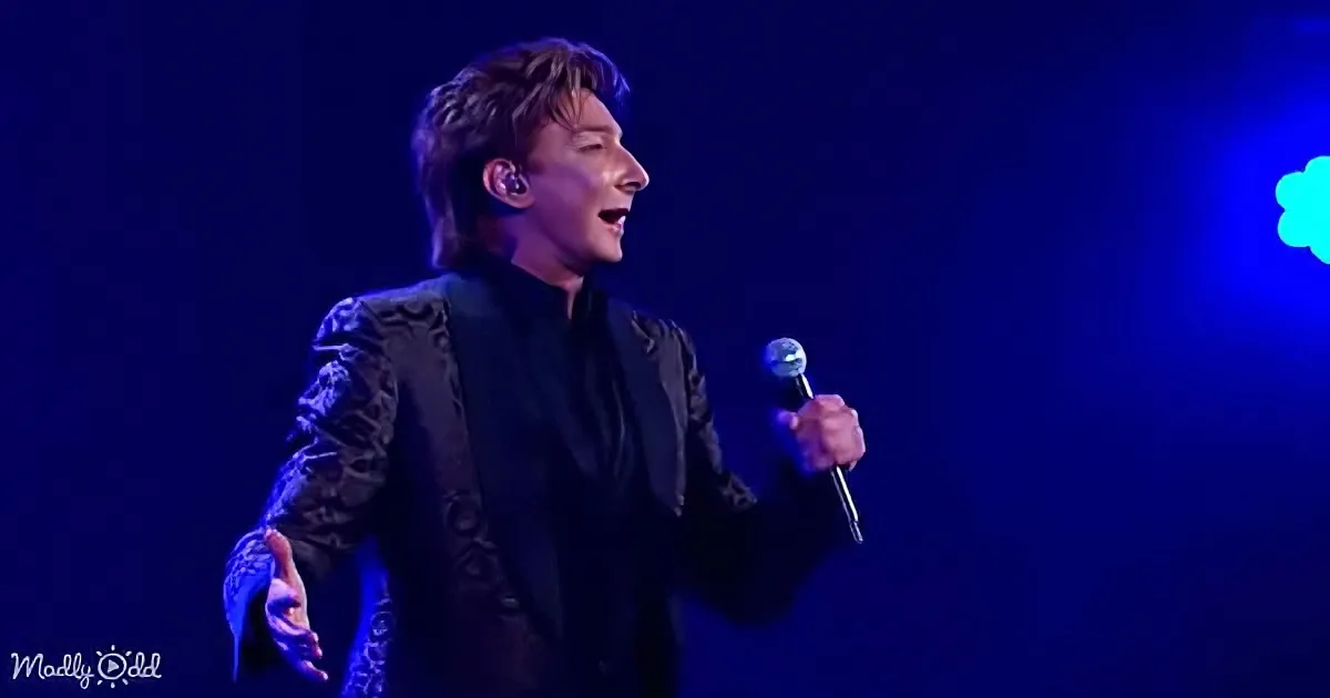 A man in a black patterned suit sings into a microphone on stage under blue lighting.