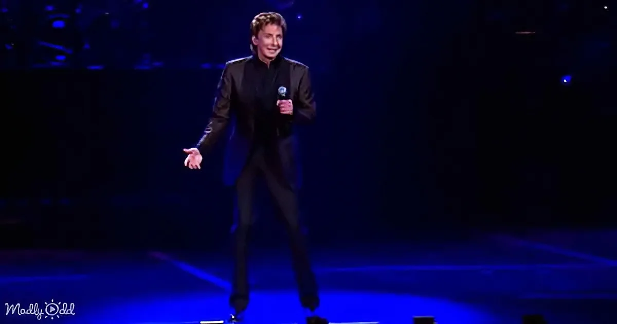 A man in a black suit holds a microphone, smiling, on a stage with blue lighting.