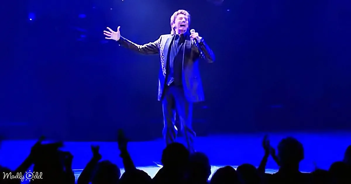 A performer in a suit sings passionately on stage under blue lighting, with an audience raising their hands in excitement.