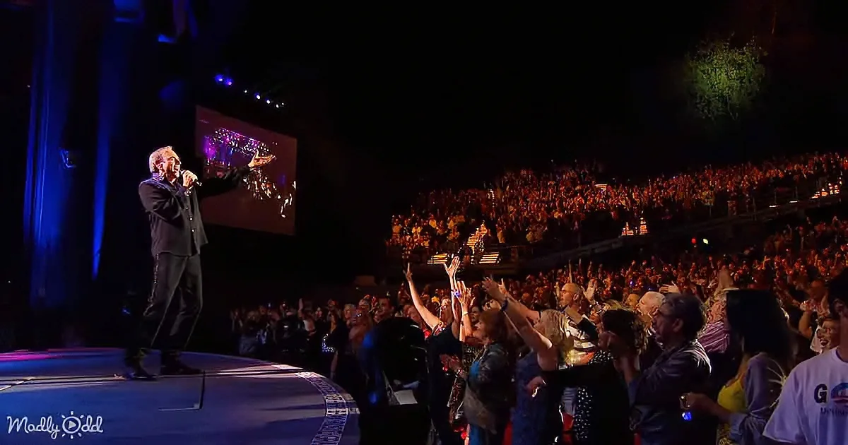 A man on stage with a microphone, surrounded by an enthusiastic crowd raising their hands.