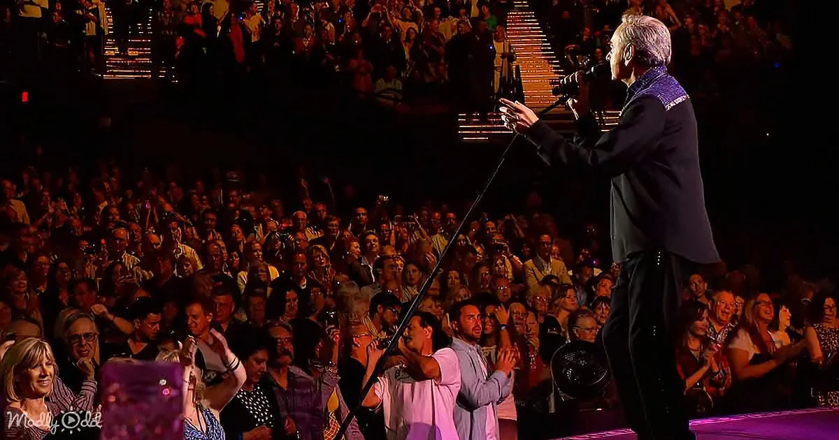 A man in a suit sings into a microphone on stage, surrounded by a cheering crowd in a large arena.