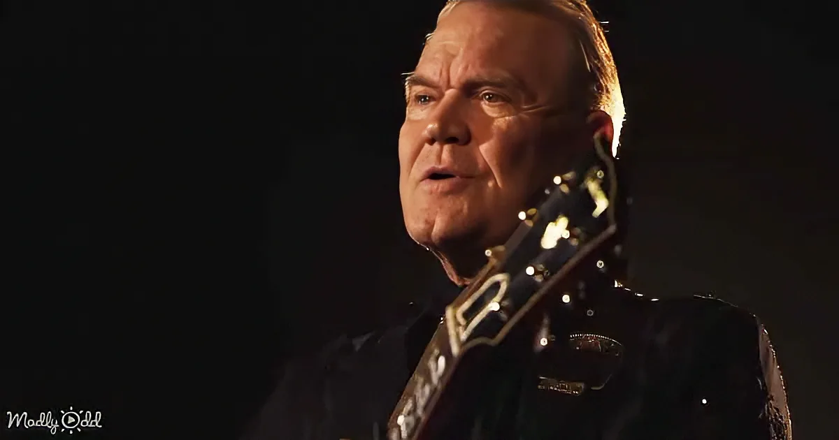 A man in a black shirt holds a guitar, illuminated by a spotlight against a dark background.