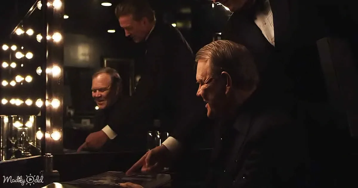A man in a suit is seated at a table, interacting with another man in a dimly lit room.