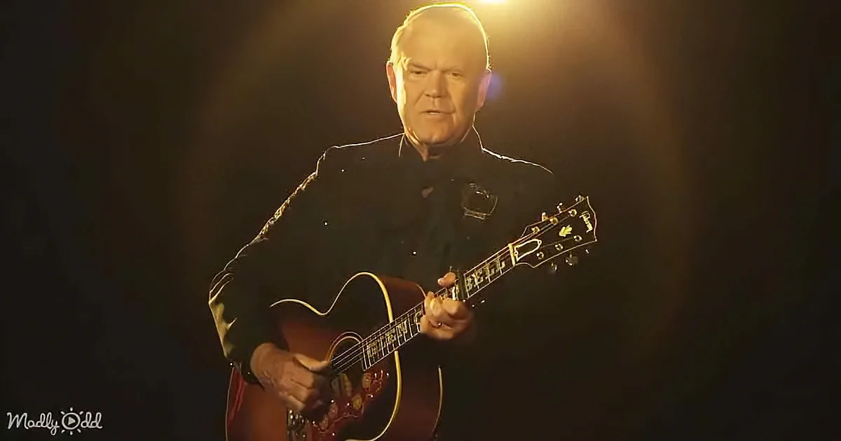 An older man in a black suit plays a red acoustic guitar under a spotlight.