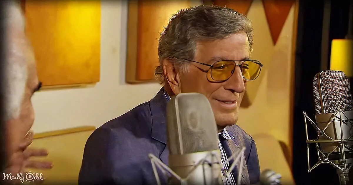 A man in a suit and sunglasses sits in a recording studio, speaking into a microphone.