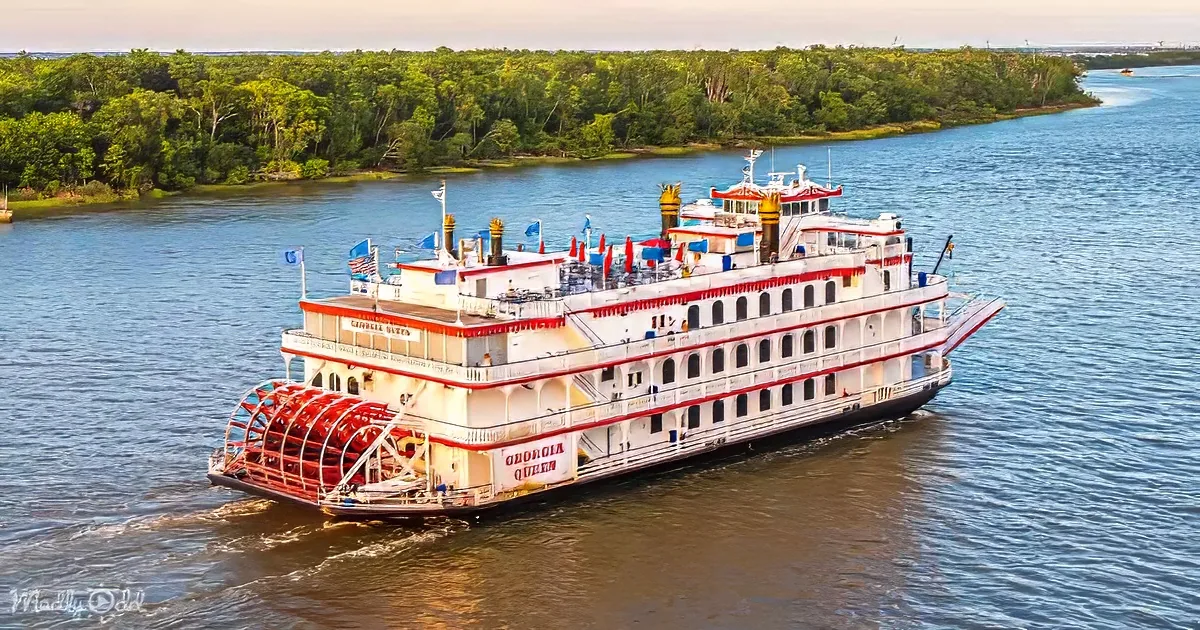 A Georgia Queen paddlewheel boat sails on a river, surrounded by lush green trees.