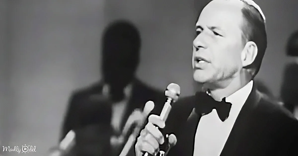 A man in a tuxedo sings into a microphone, with blurred figures in the background.