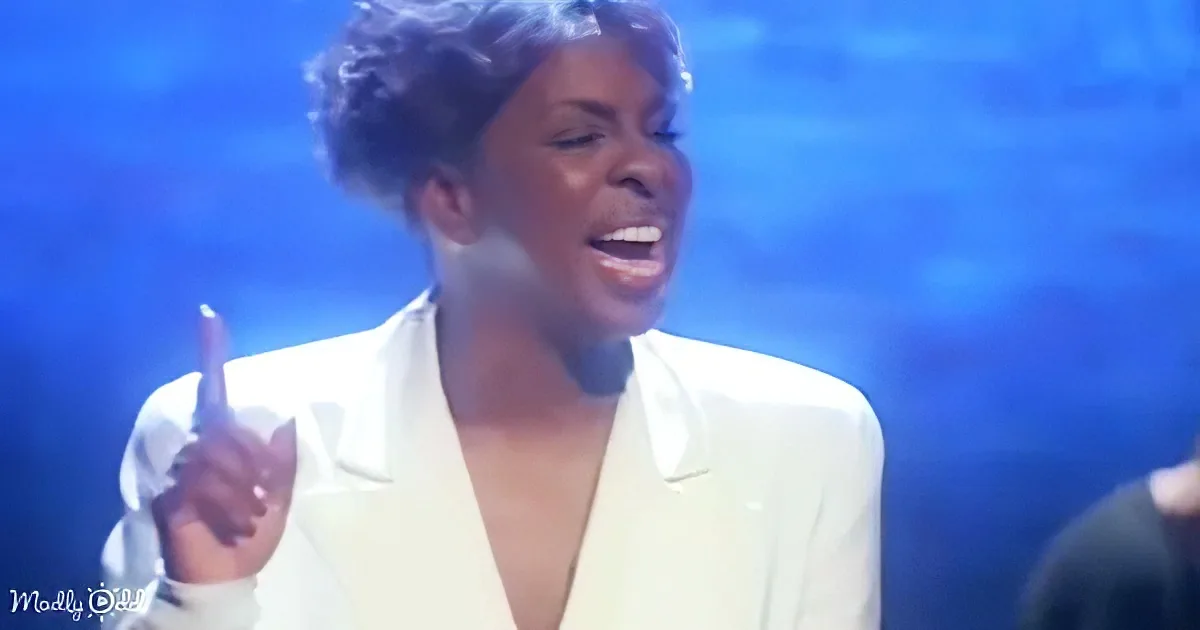 A person in a white blazer gestures with their right hand while singing or speaking.