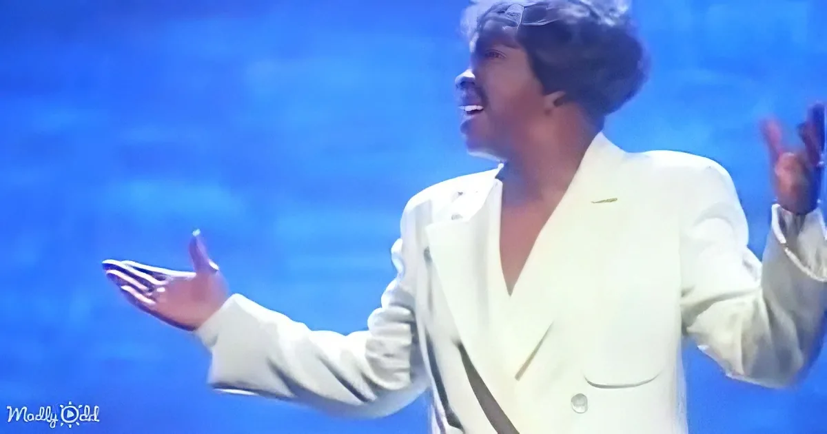A person in a white suit gestures dramatically against a blue background.