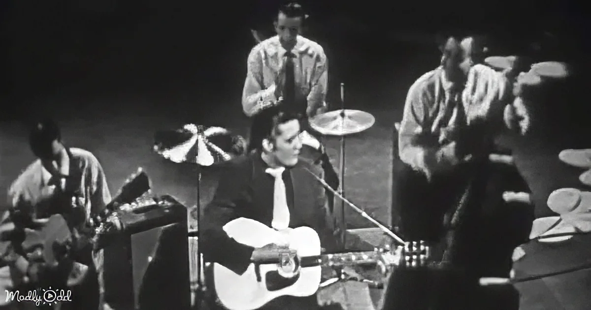 Elvis Presley plays guitar on stage with a band, wearing a suit and tie.