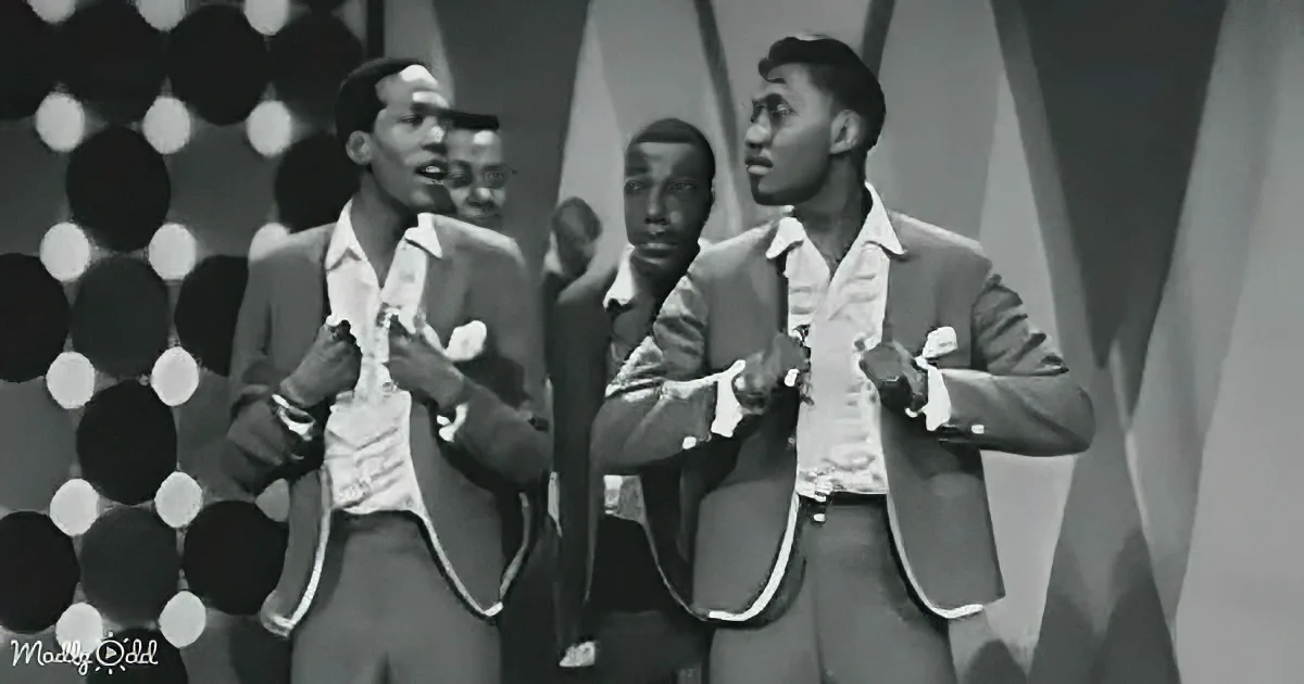 Four men in suits sing on stage, one holding a microphone, against a backdrop of circular lights.