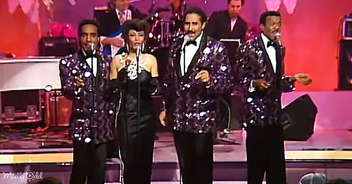 A group of performers in shiny jackets and a woman in a black dress sing on stage with a band behind them.