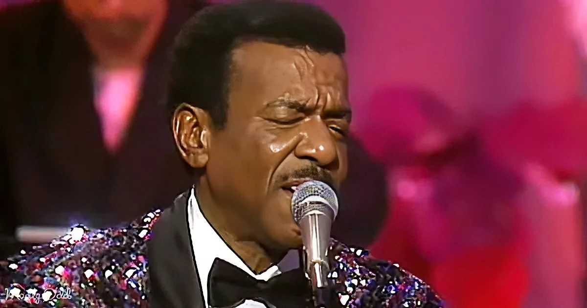 A man in a glittery suit sings into a microphone on stage.