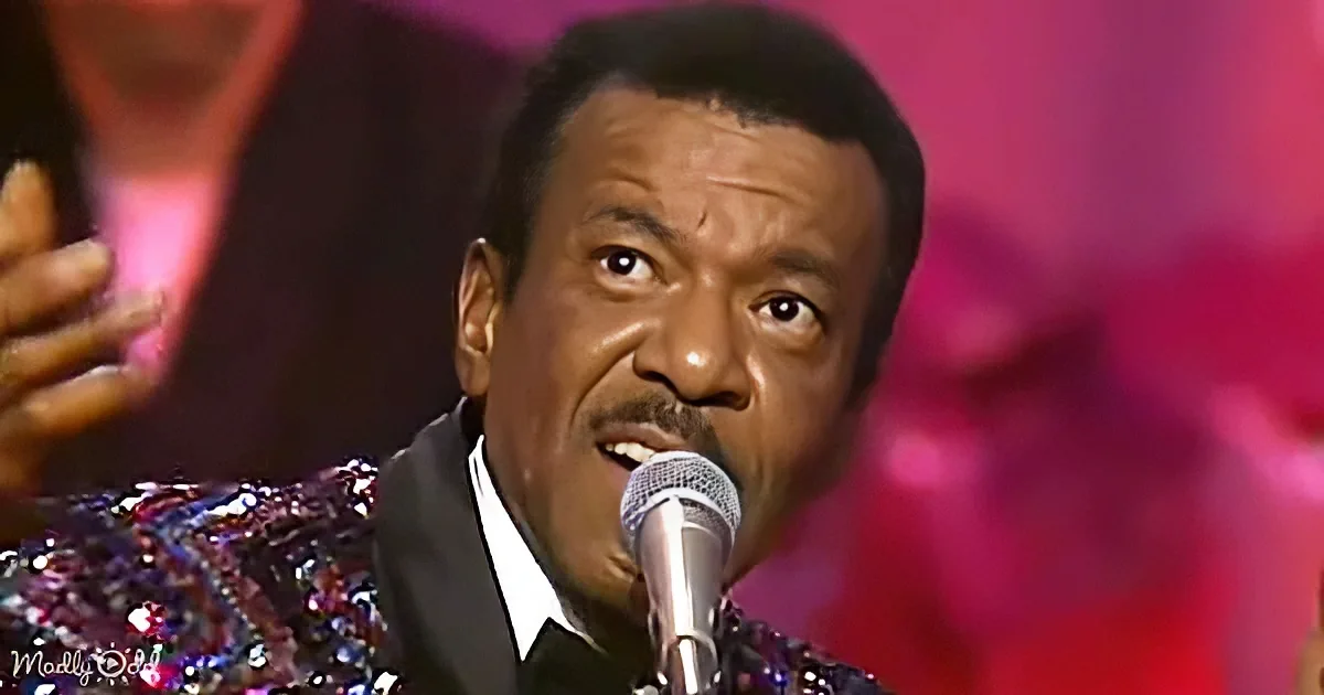A man in a sequined jacket sings into a microphone, with a blurred background of red and pink hues.
