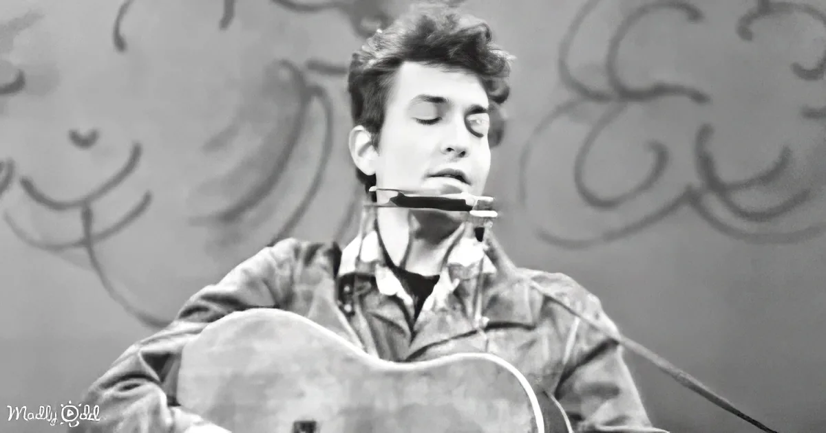 A man plays guitar and harmonica, wearing a jacket, with a graffiti-covered wall behind him.