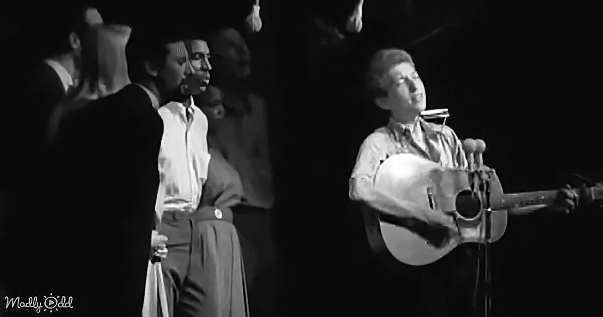 A man plays guitar on stage, singing, with a group of musicians in the background.