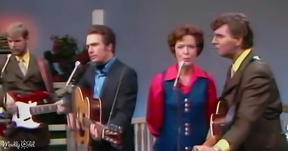 A group of musicians, including a woman in a red jacket, perform on stage with guitars and microphones.