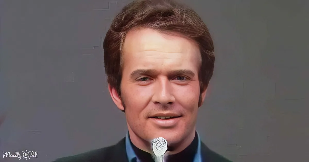 A man with short brown hair, wearing a dark suit and blue shirt, smiles near a microphone against a gray background.