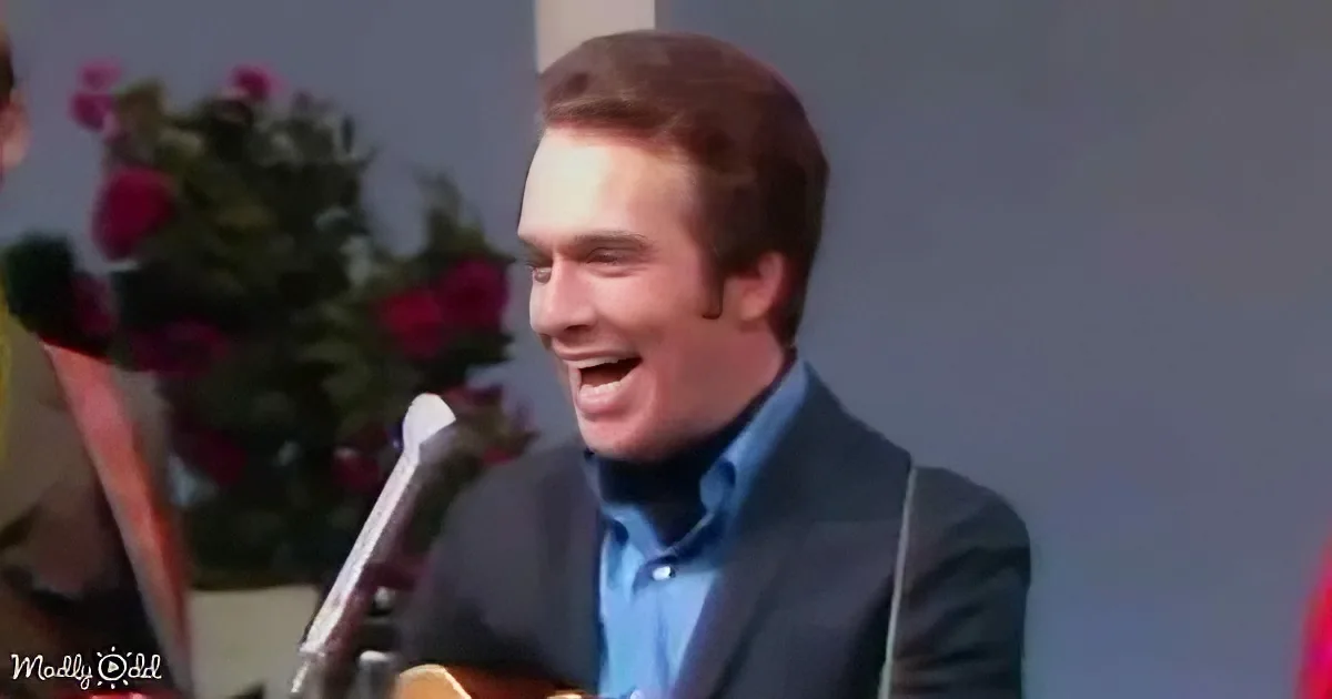 A man in a suit and blue shirt smiles while playing a guitar, with flowers in the background.