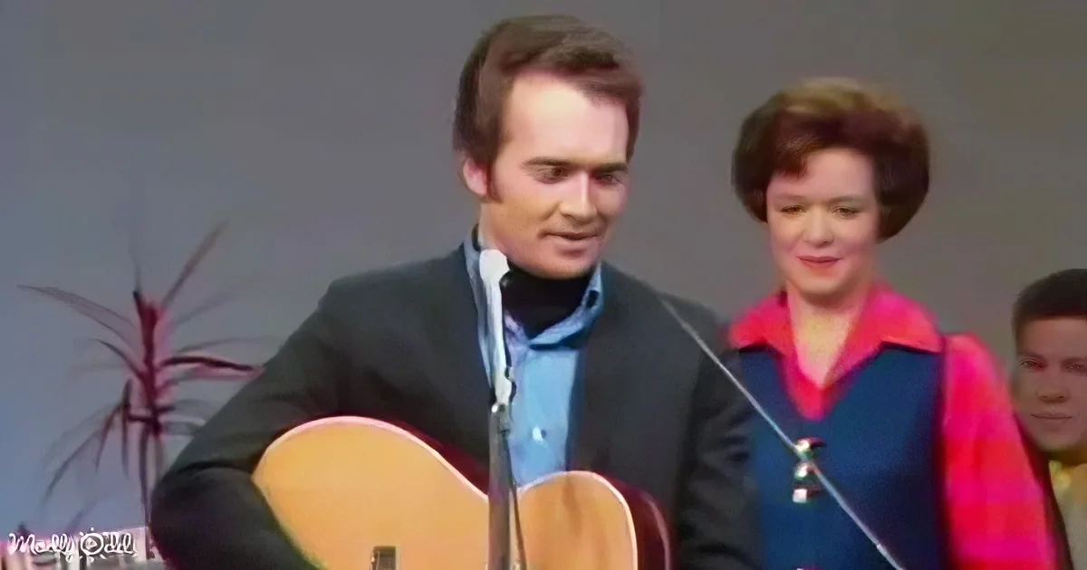 A man in a suit plays guitar on stage, with a woman in a colorful outfit playing violin behind him.