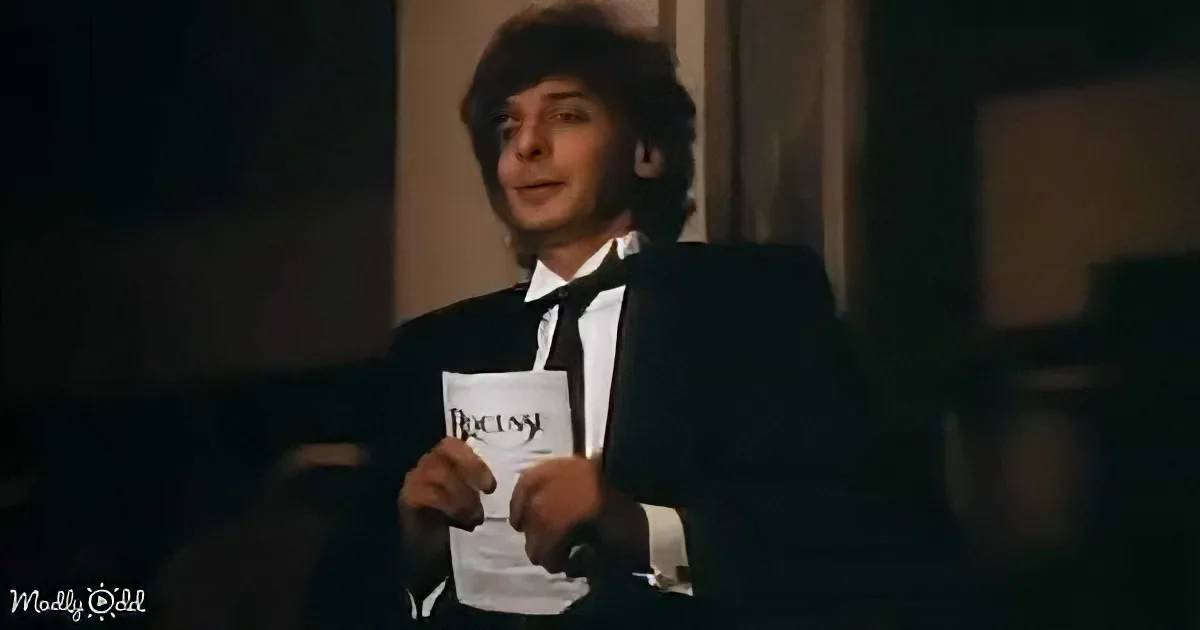 A man in a tuxedo holds a white envelope with "Puccini" written on it, smiling.