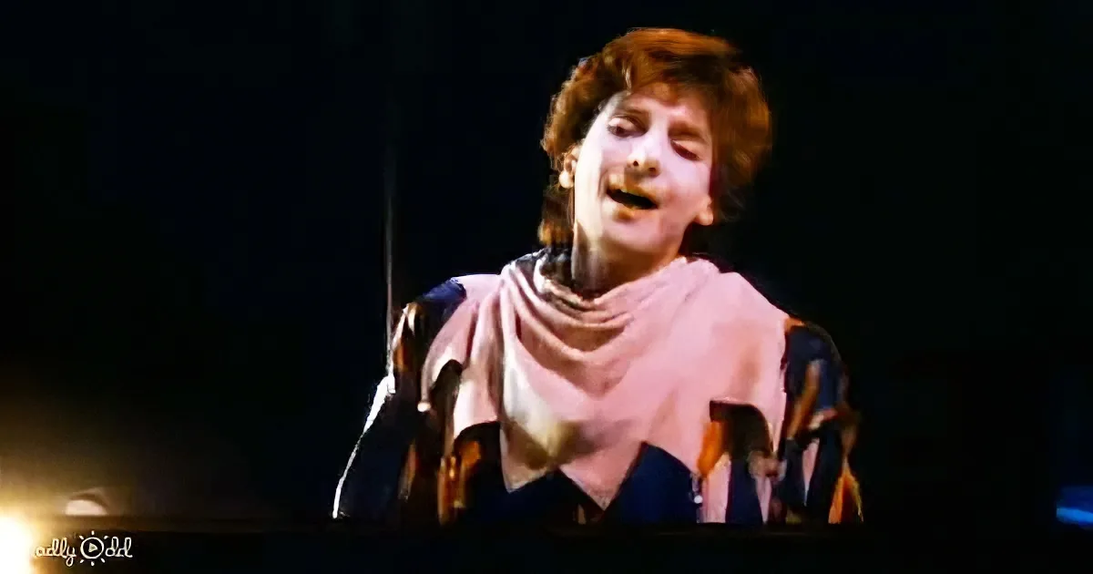 A person in a colorful outfit with a pink scarf is singing on stage, illuminated by stage lights.