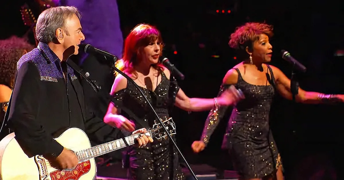 A man plays guitar while two women sing on stage with microphones.