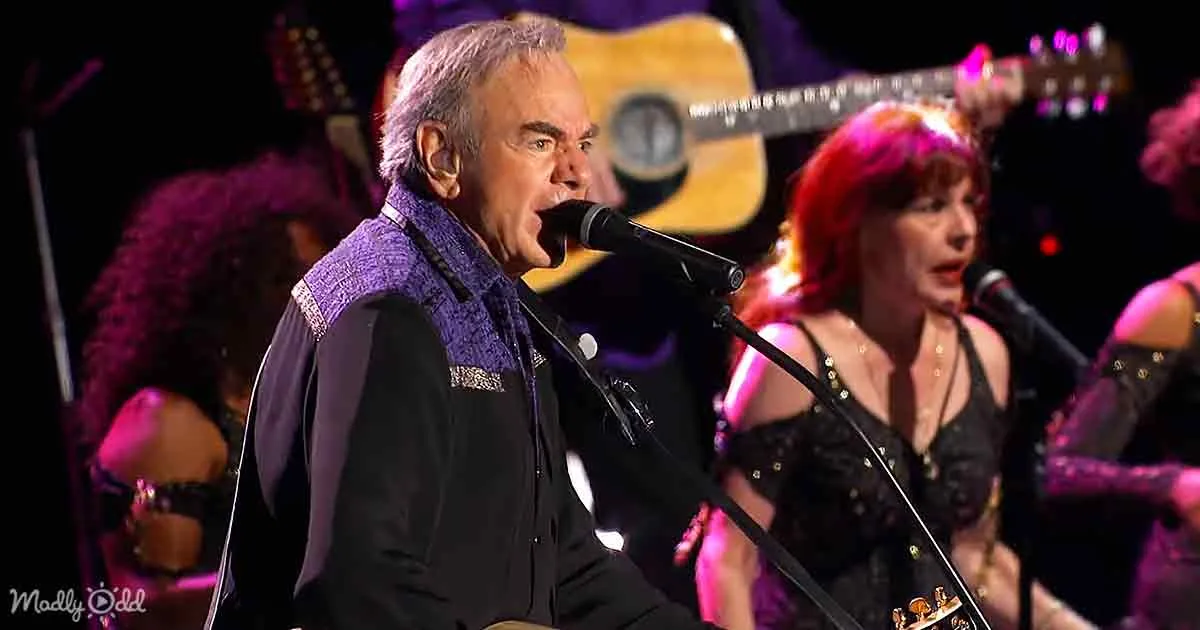 A man in a purple vest sings into a microphone on stage with a band.