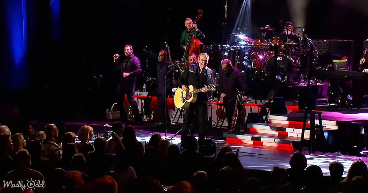 A man in a suit plays guitar on stage with a band and audience in the foreground.