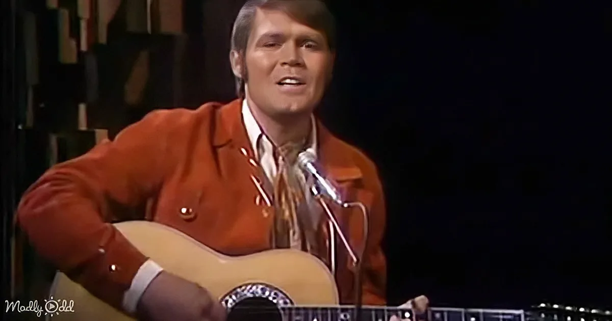 A man in a red jacket plays an acoustic guitar on stage, singing into a microphone.