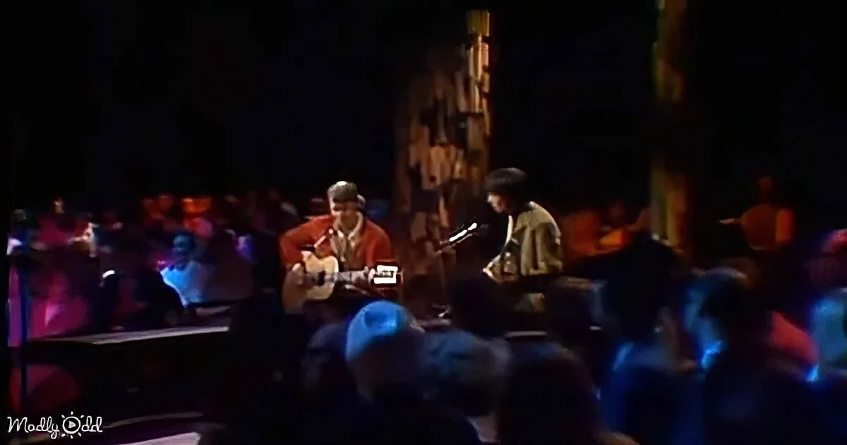 Two musicians play acoustic guitars on stage, surrounded by an audience in a dimly lit venue.