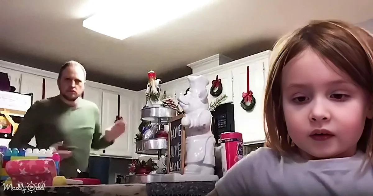 A child sits in the foreground, while a man in a green sweater stands in a kitchen decorated for Christmas.