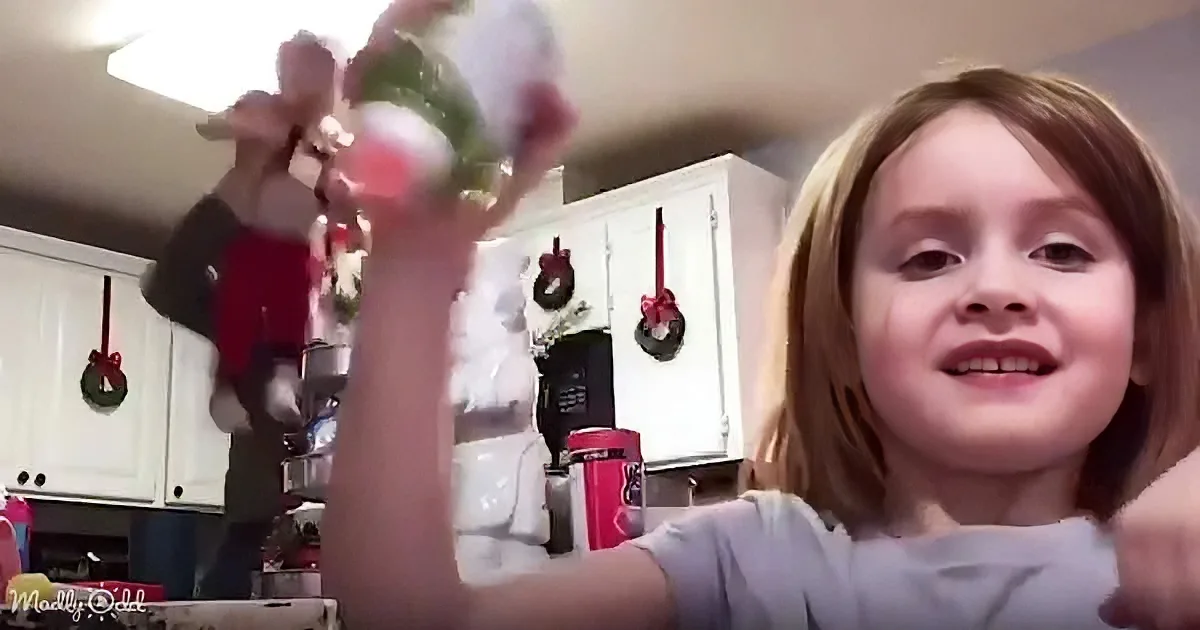 A young girl with a toothless smile holds up a Christmas ornament in a kitchen decorated with wreaths.