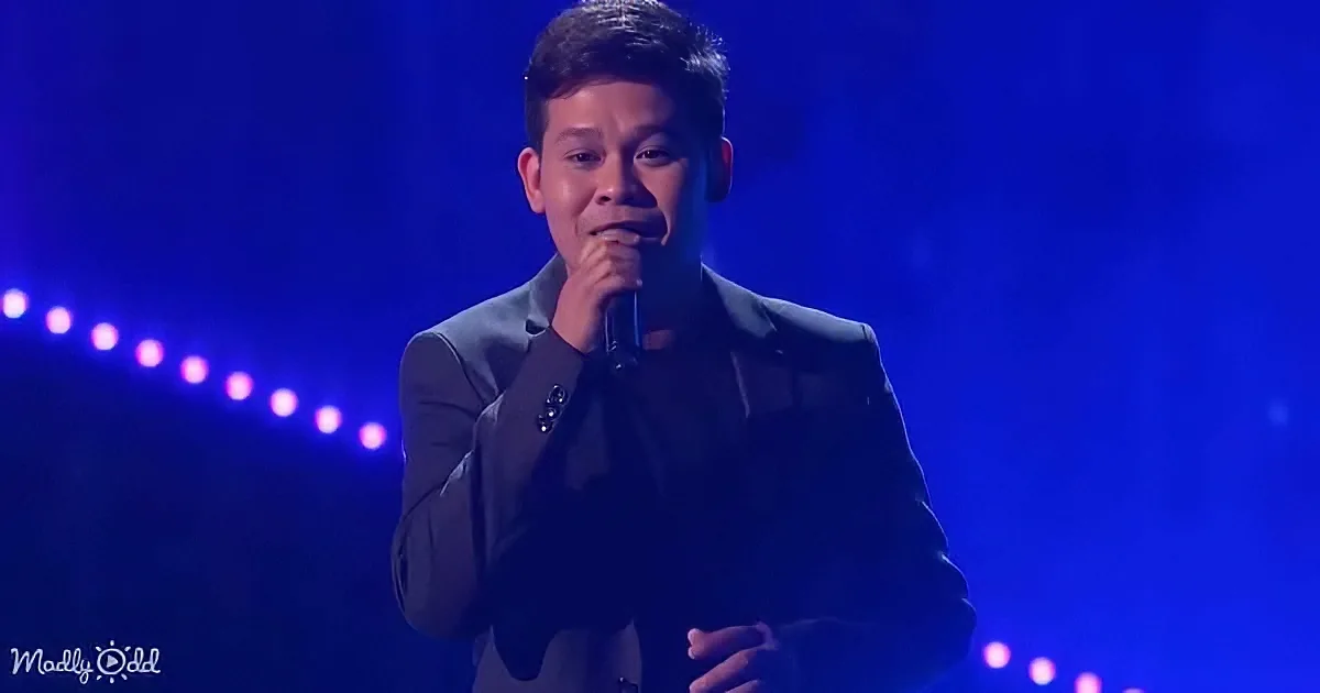 A man in a suit sings into a microphone on a stage with blue lighting.