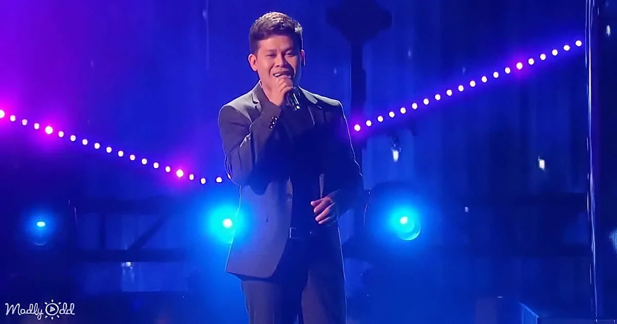 A man in a suit sings on stage under purple and blue lights.