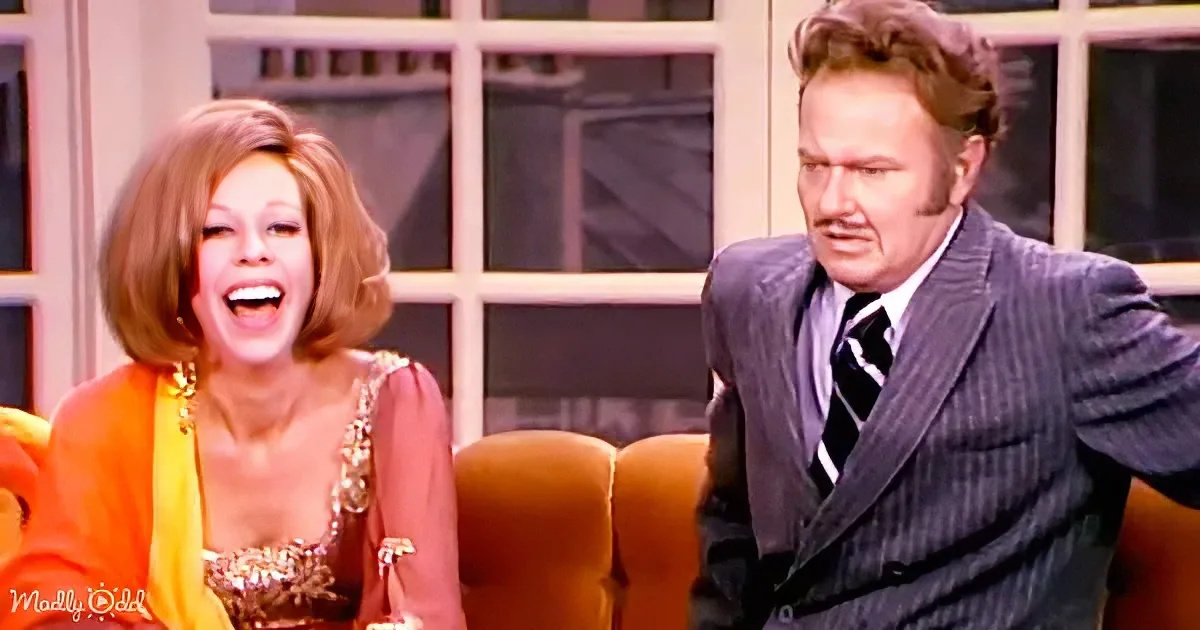 Woman in orange and gold outfit laughs, man in pinstripe suit leans back, both seated on a couch.