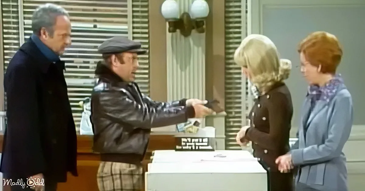 A man in a leather jacket points a finger, while others observe in a room with a counter and lamp.