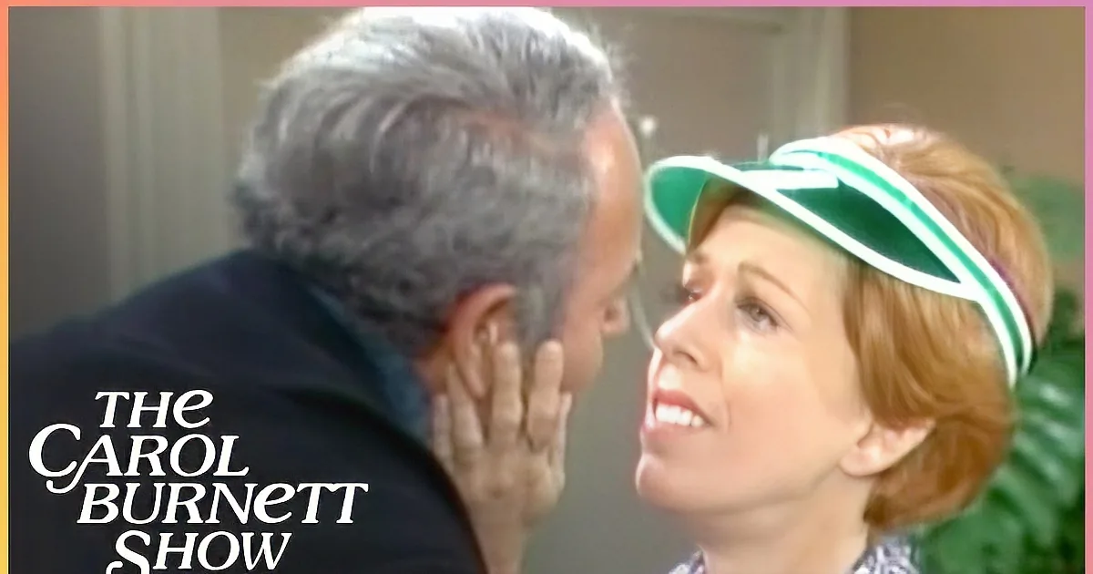 A woman in a green visor and patterned top looks at a man in a dark jacket, both in a room with a plant.