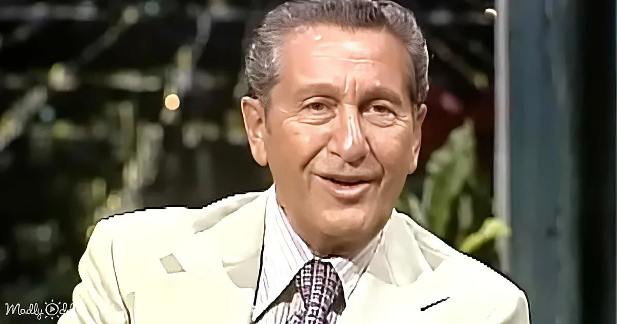 A man in a white suit and patterned tie smiles, with blurred greenery and a structure in the background.