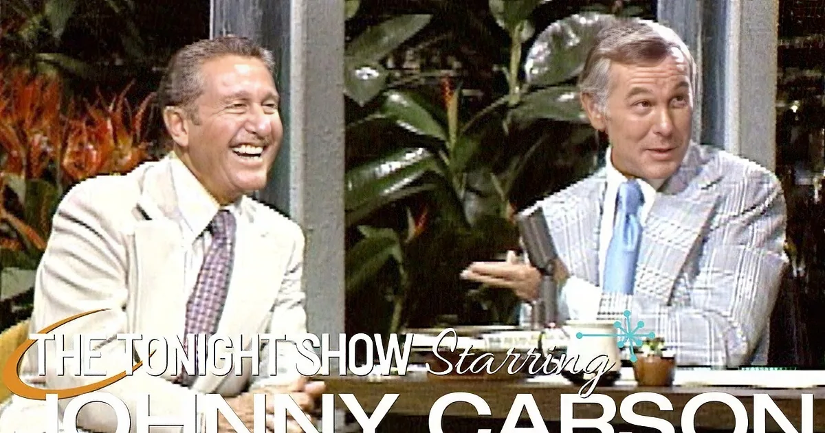 Two men in suits sit at a desk, one laughing, the other speaking, with plants in the background.