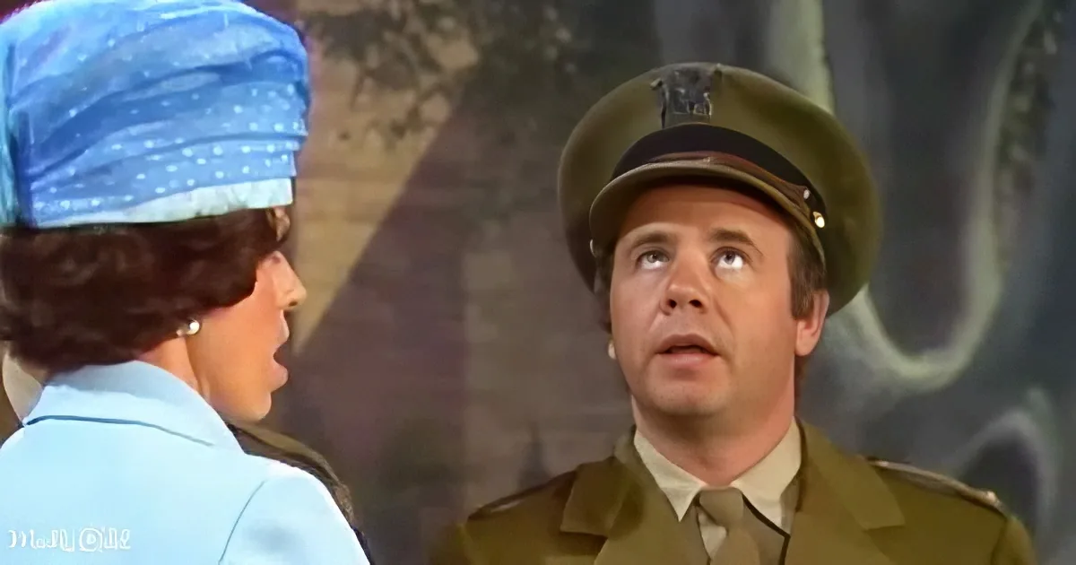 A man in a military uniform looks up while a woman in a blue outfit speaks to him.