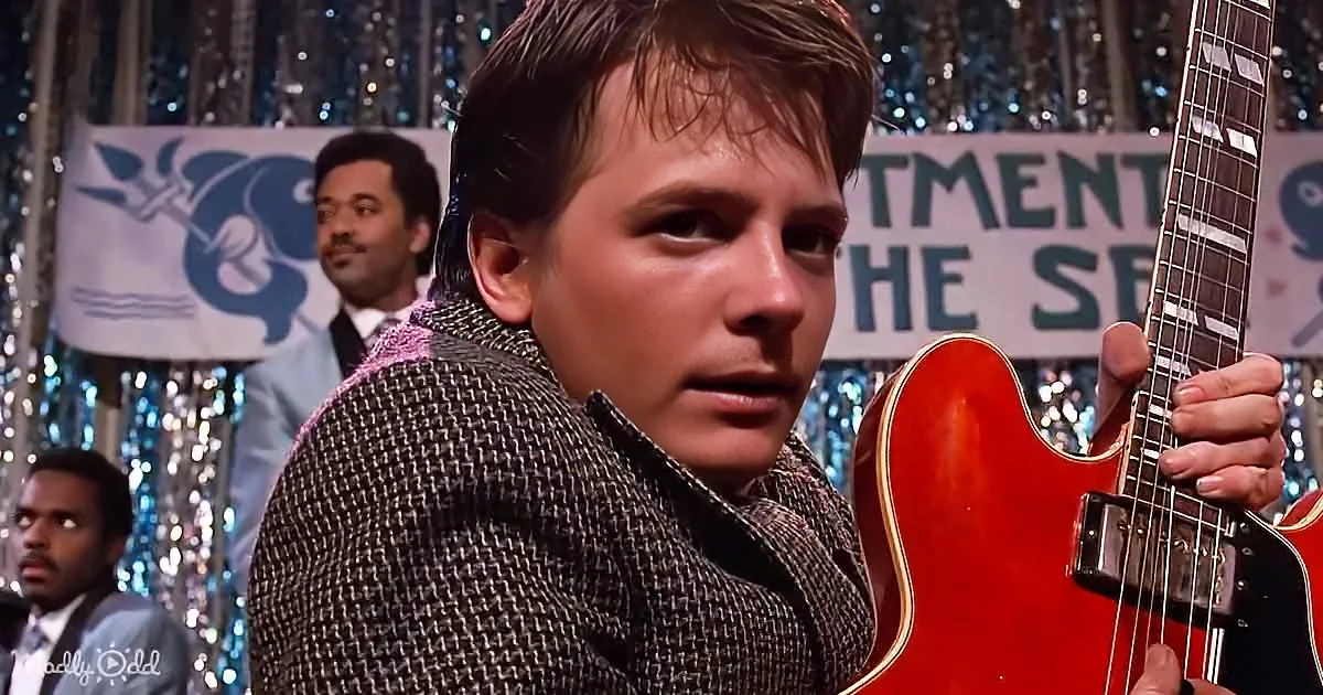 A man in a patterned suit holds a red electric guitar, with a shiny backdrop and other musicians in the background.