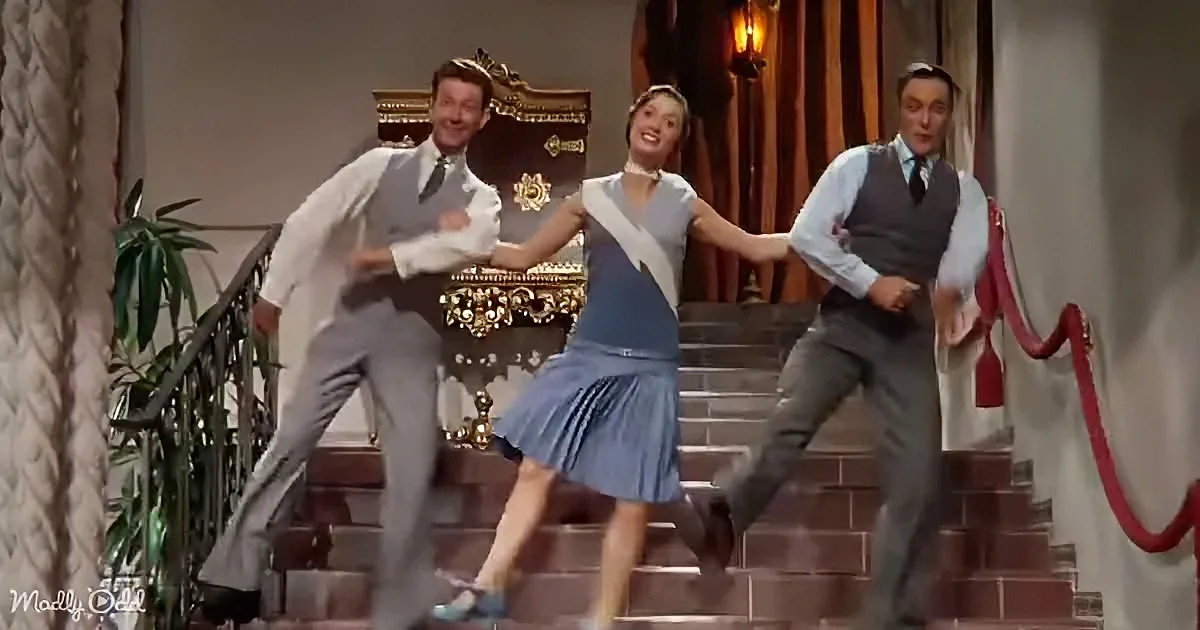 A woman in a blue dress dances up stairs with two men in suits, creating a lively scene.